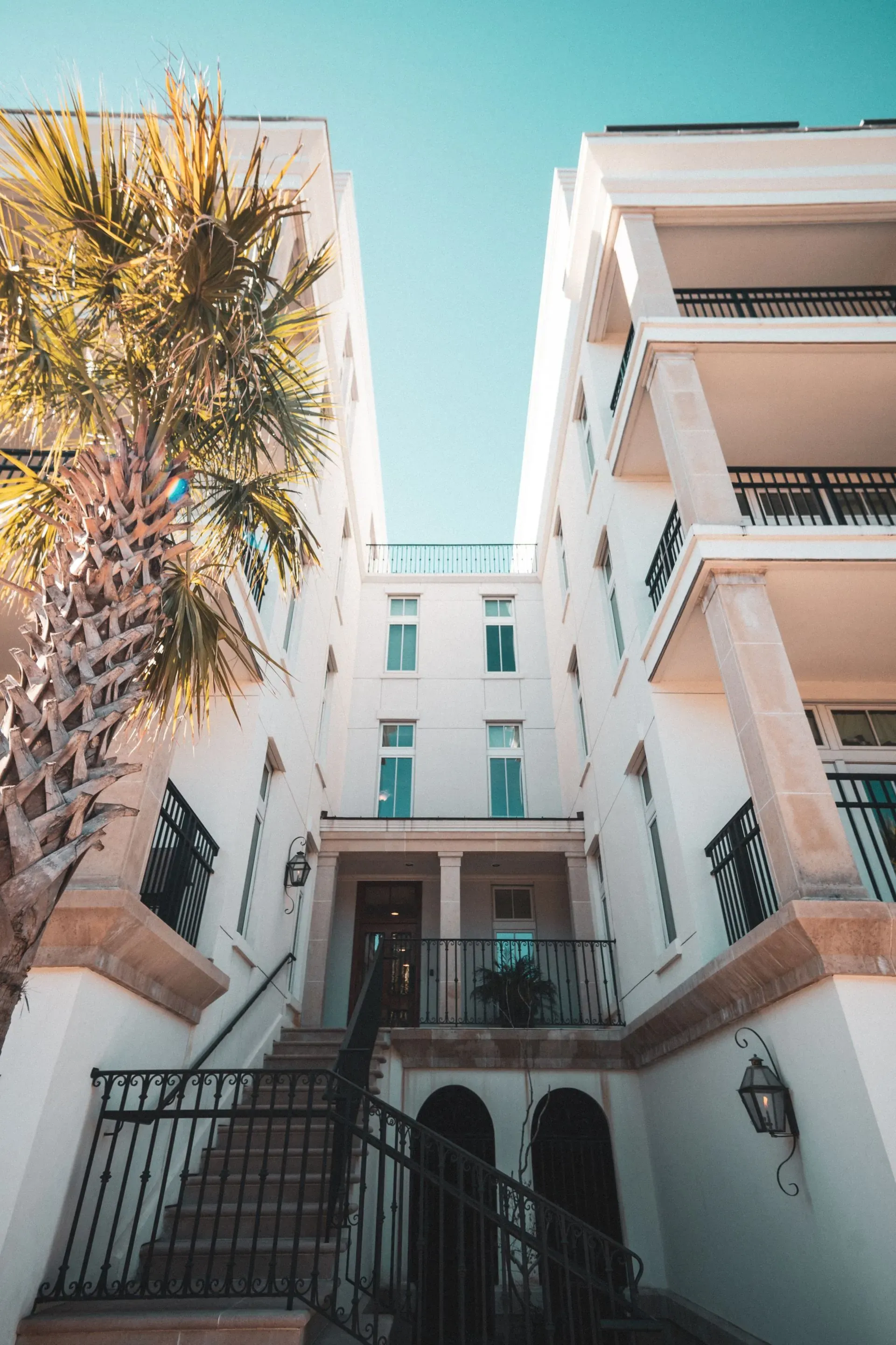 Coastal apartment courtyard with balconies, palm tree, and bright blue sky.
